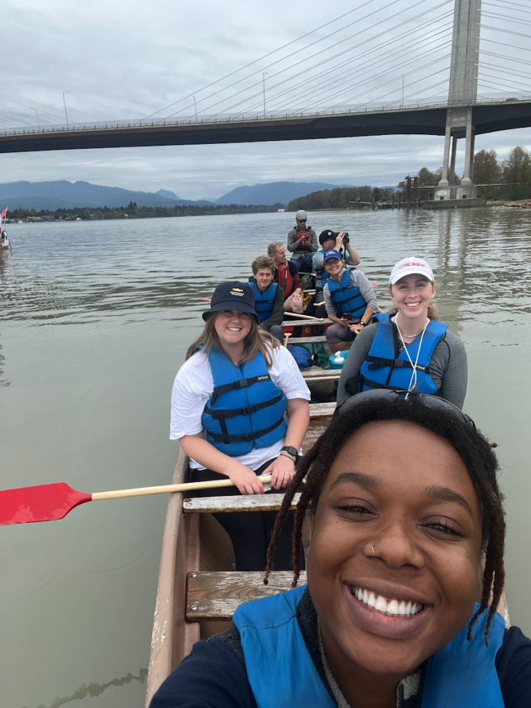 Eight people sitting and smiling in a canoe with the Golden Ears Bridge and mountains in the background